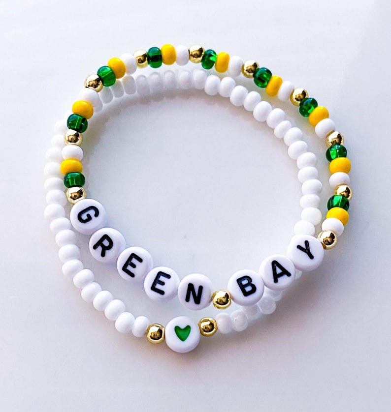 May include: Two white beaded bracelets. One bracelet spells out "GREEN BAY" in black letters on white beads. The other bracelet has green, yellow, and gold beads. A green heart bead is also visible.