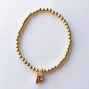 May include: Gold beaded bracelet with a pizza slice charm. The bracelet features small, round gold beads strung together, forming a circular shape. A small, detailed pizza slice charm hangs from the bracelet, adding a playful touch. The bracelet is set against a white background.