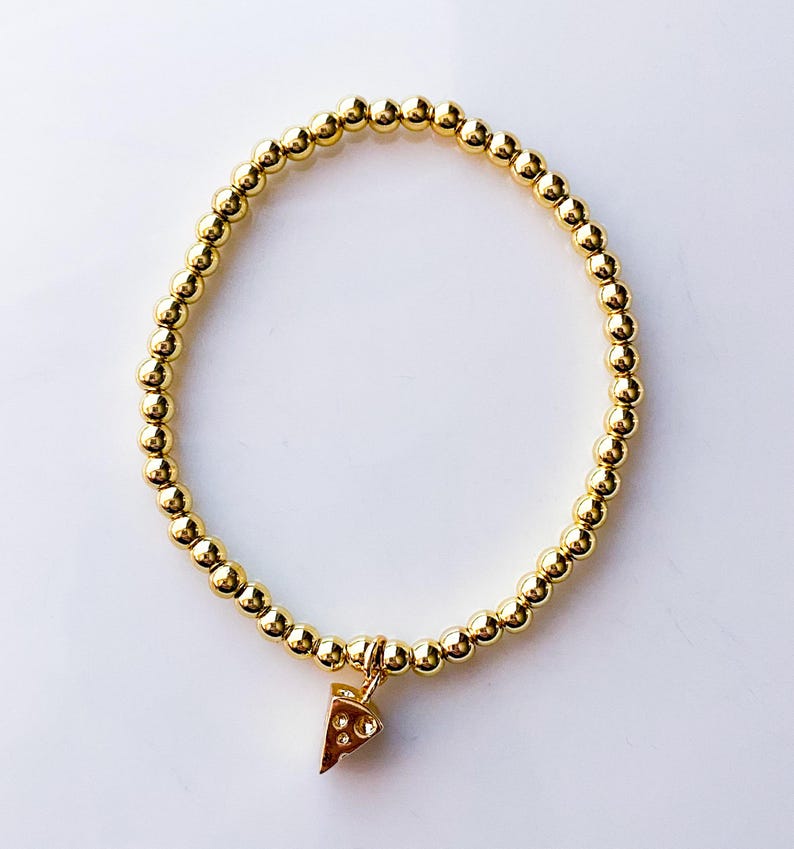 May include: Gold beaded bracelet with a pizza slice charm. The bracelet features small, round gold beads strung together, forming a circular shape. A small, detailed pizza slice charm hangs from the bracelet, adding a playful touch. The bracelet is set against a white background.