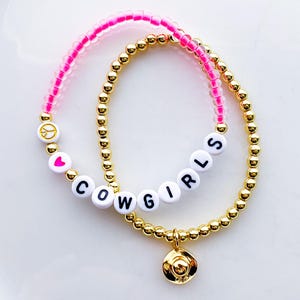 Puede incluir: Dos pulseras de cuentas. Una es dorada con un peque&ntilde;o dije de sombrero de vaquero dorado. La otra es rosa con cuentas de letras blancas que deletrean "COWGIRLS", un signo de paz y un coraz&oacute;n.