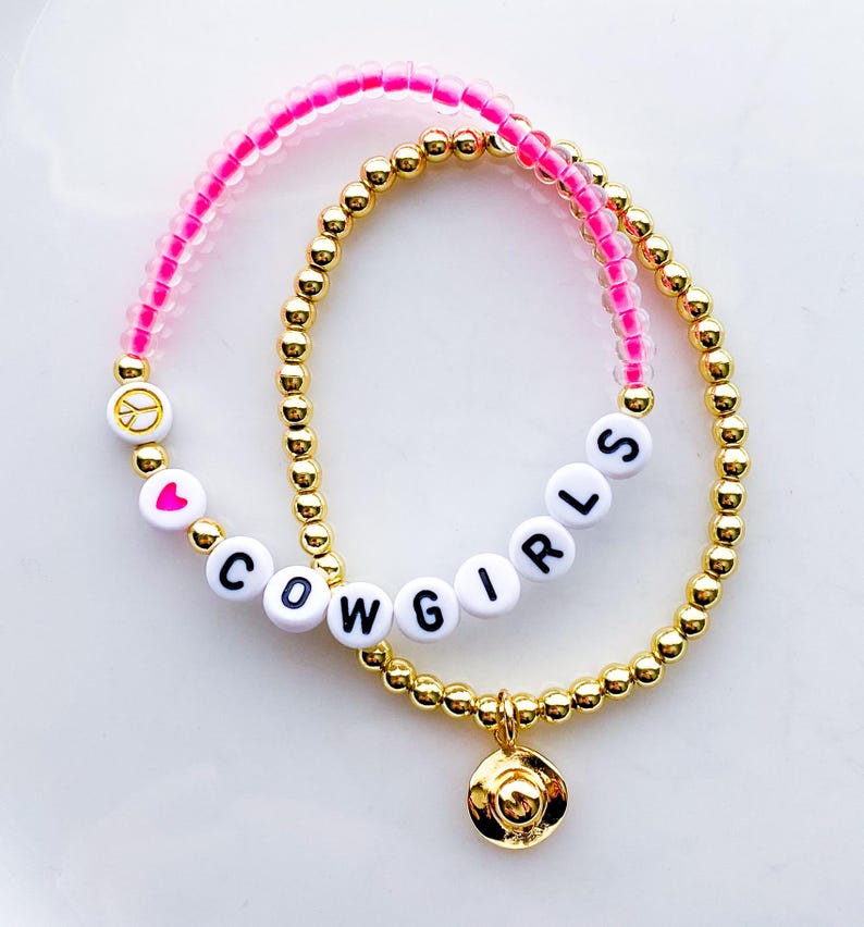 Puede incluir: Dos pulseras de cuentas. Una es dorada con un peque&ntilde;o dije de sombrero de vaquero dorado. La otra es rosa con cuentas de letras blancas que deletrean "COWGIRLS", un signo de paz y un coraz&oacute;n.
