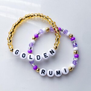 Pode incluir: Duas pulseiras de contas. Uma &eacute; dourada com a palavra "GOLDEN" escrita com contas de letras brancas. A outra pulseira &eacute; roxa, branca e dourada com a palavra "RUMI" escrita com contas de letras brancas.