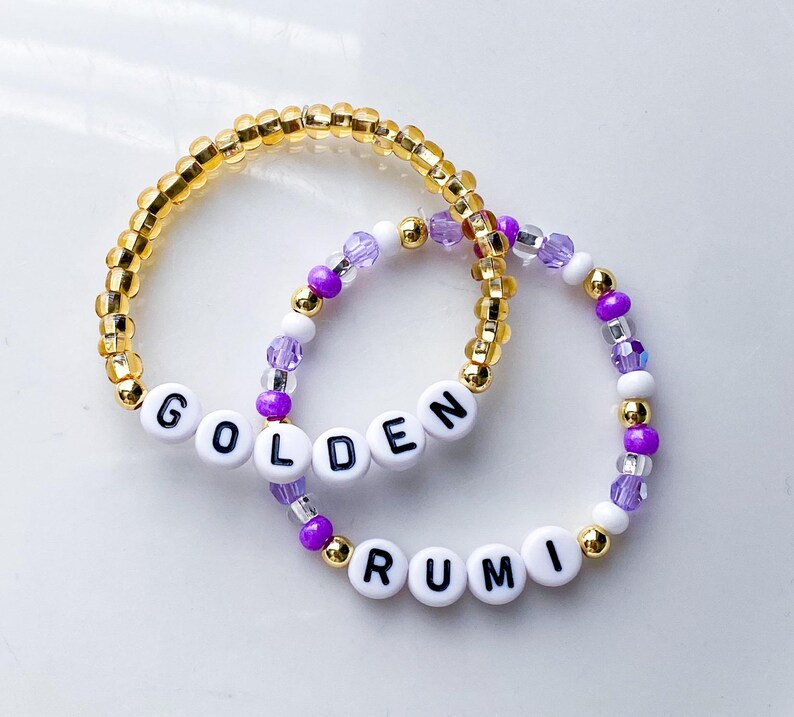 Pode incluir: Duas pulseiras de contas. Uma &eacute; dourada com a palavra "GOLDEN" escrita com contas de letras brancas. A outra pulseira &eacute; roxa, branca e dourada com a palavra "RUMI" escrita com contas de letras brancas.