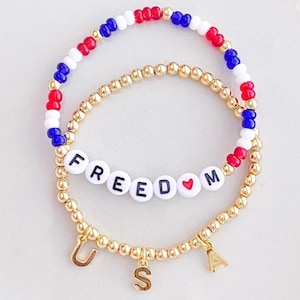 May include: Two beaded bracelets. One bracelet features red, white, and blue beads. The other bracelet has gold beads with the word "FREEDOM" spelled out in white letter beads and a heart symbol. Gold letter charms spell "USA".