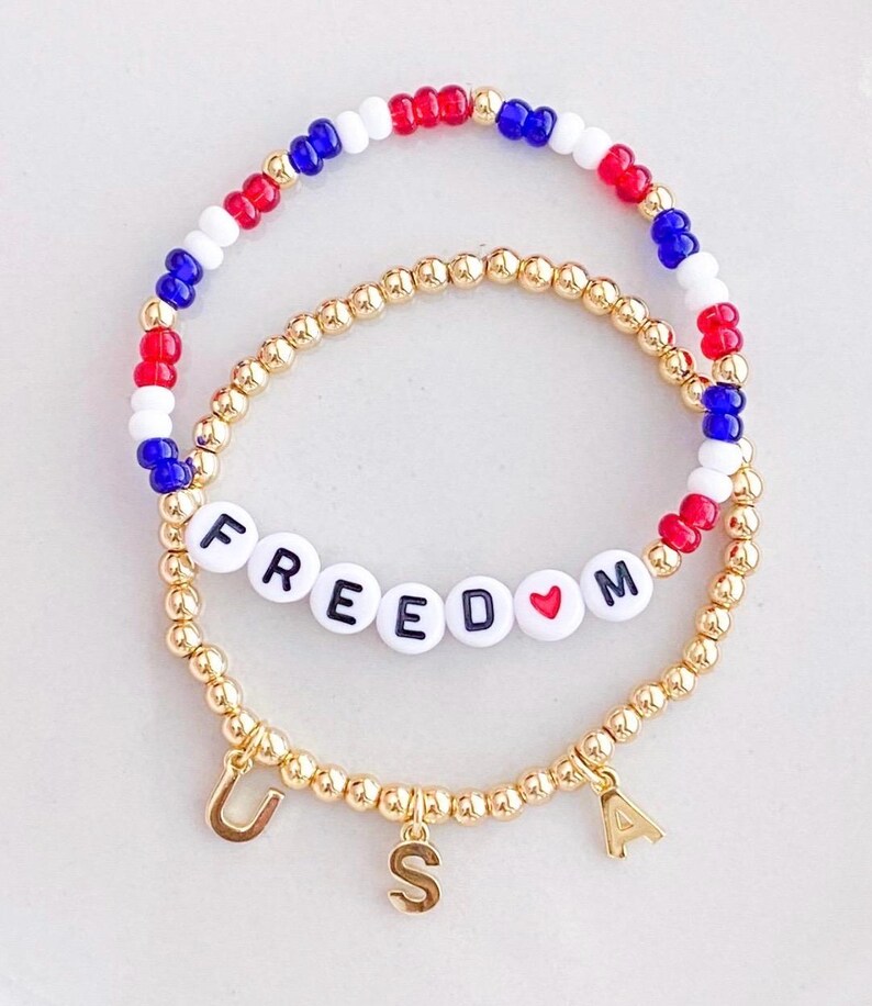 May include: Two beaded bracelets. One bracelet features red, white, and blue beads. The other bracelet has gold beads with the word "FREEDOM" spelled out in white letter beads and a heart symbol. Gold letter charms spell "USA".