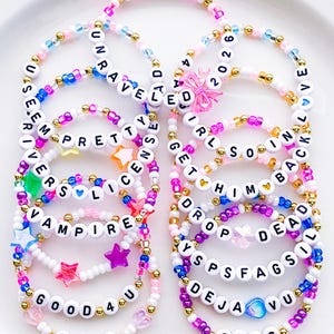 May include: A collection of beaded bracelets in various colors, including pink, blue, and white. Each bracelet features white letter beads spelling out words and phrases like "Vampire" and "Deja Vu". Some bracelets have star and heart charms.
