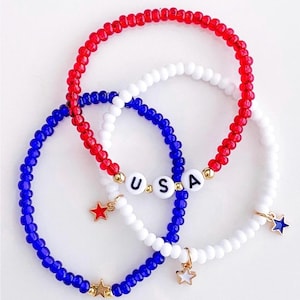 May include: Three beaded bracelets in red, white, and blue, with the letters "USA" spelled out on one. Each bracelet has a star charm. The bracelets are a patriotic accessory.