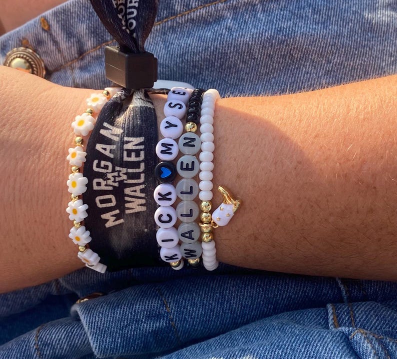 K&ouml;nnte beinhalten: Eine Sammlung von Armb&auml;ndern mit Perlen und ein schwarzes Armband mit wei&szlig;em Text, der "MORGAN WALLEN" lautet. Die Armb&auml;nder umfassen bl&uuml;tenf&ouml;rmige Perlen, Buchstabenperlen mit der Aufschrift "KICK MYSELF WALLEN" und ein wei&szlig;es Armband mit einem goldenen Anh&auml;nger.