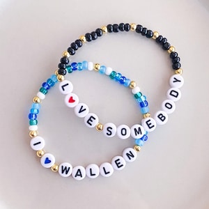 K&ouml;nnte beinhalten: Zwei Armb&auml;nder mit Perlen, auf denen die W&ouml;rter "I LOVE SOMEBODY WALLEN" mit wei&szlig;en Buchstabenperlen geschrieben sind. Die Armb&auml;nder haben eine Mischung aus blauen, schwarzen und goldenen Perlen. Ein Armband hat eine rote Herzperle und das andere eine blaue Herzperle.