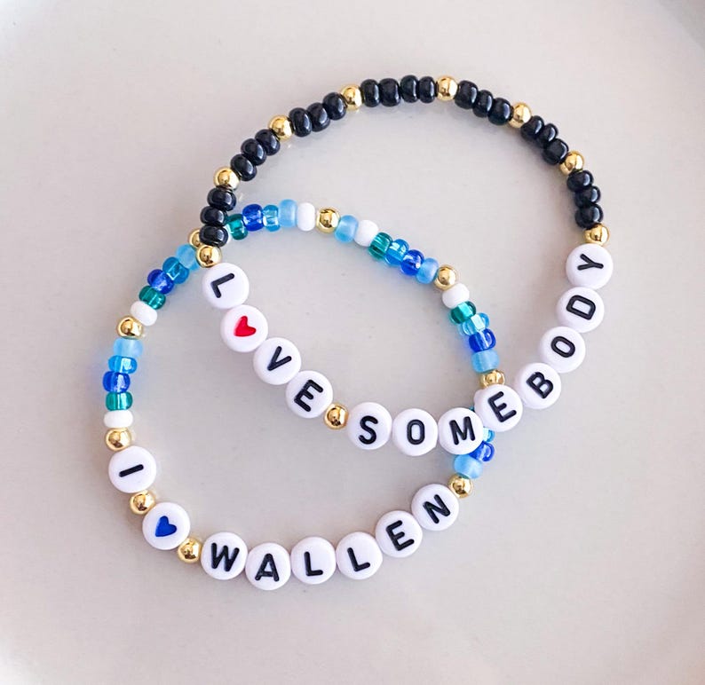 K&ouml;nnte beinhalten: Zwei Armb&auml;nder mit Perlen, auf denen die W&ouml;rter "I LOVE SOMEBODY WALLEN" mit wei&szlig;en Buchstabenperlen geschrieben sind. Die Armb&auml;nder haben eine Mischung aus blauen, schwarzen und goldenen Perlen. Ein Armband hat eine rote Herzperle und das andere eine blaue Herzperle.