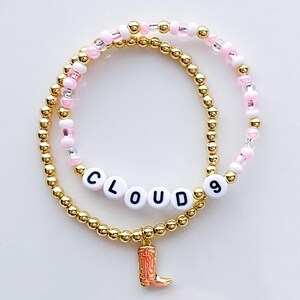 Puede incluir: Dos pulseras de cuentas. Una es dorada con la palabra "CLOUD 9" escrita con cuentas de letras blancas. La otra pulsera es rosa y blanca con cuentas doradas. Un peque&ntilde;o dije de bota naranja cuelga de la pulsera inferior.