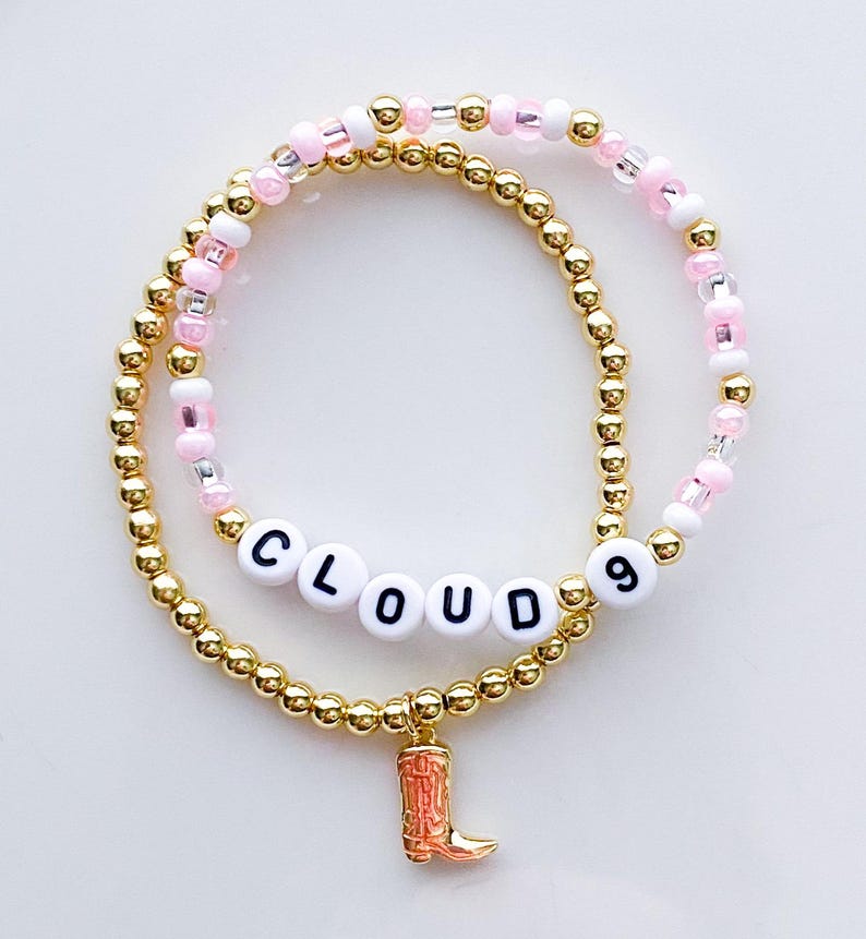 Puede incluir: Dos pulseras de cuentas. Una es dorada con la palabra "CLOUD 9" escrita con cuentas de letras blancas. La otra pulsera es rosa y blanca con cuentas doradas. Un peque&ntilde;o dije de bota naranja cuelga de la pulsera inferior.