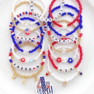 May include: A collection of beaded bracelets in red, white, and blue, with gold accents. Some bracelets spell out "USA," "FREEDOM," "AMERICA," and "WHITE & BLUE." Charms include stars and a small American flag. A sticker reads "4th of July."