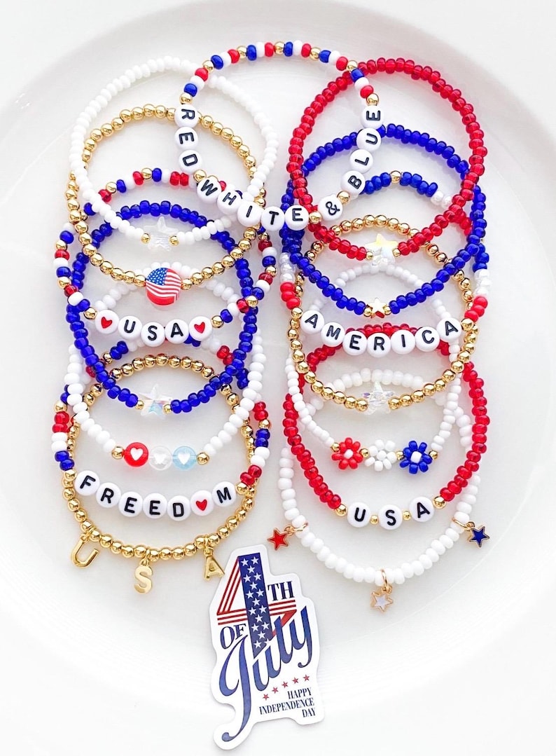 May include: A collection of beaded bracelets in red, white, and blue, with gold accents. Some bracelets spell out "USA," "FREEDOM," "AMERICA," and "WHITE & BLUE." Charms include stars and a small American flag. A sticker reads "4th of July."