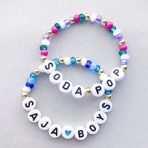 Pode incluir: Duas pulseiras de contas. Uma pulseira diz "SODA POP" com contas de letras brancas e detalhes dourados. A outra pulseira diz "SAJA BOYS" com contas de letras brancas e um cora&ccedil;&atilde;o azul. As pulseiras t&ecirc;m contas rosa, verdes, azuis e brancas.