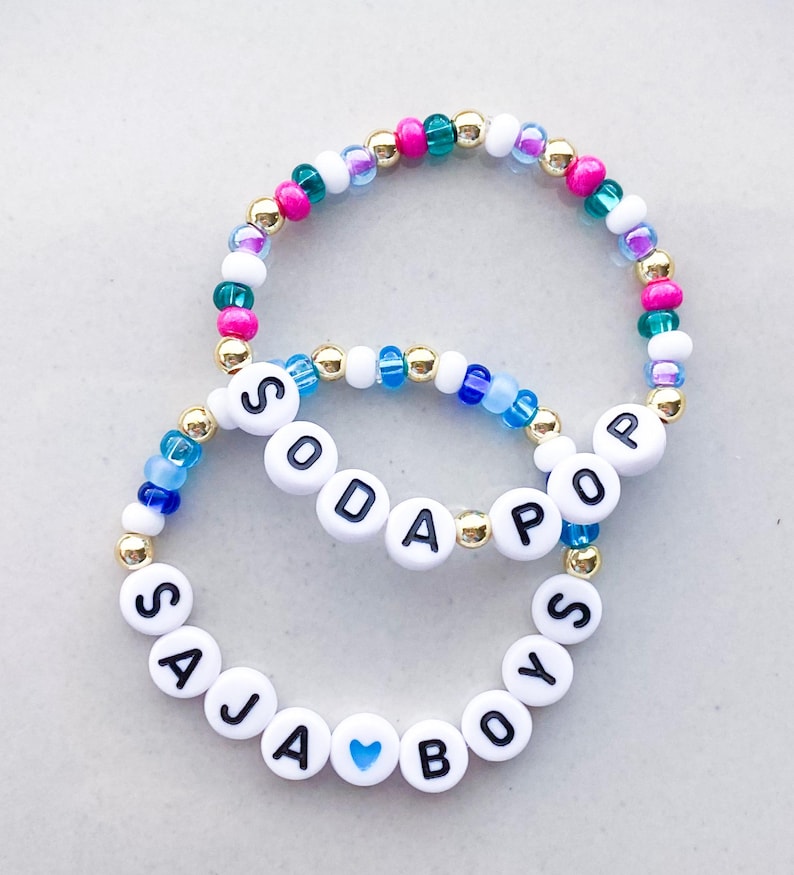 Pode incluir: Duas pulseiras de contas. Uma pulseira diz "SODA POP" com contas de letras brancas e detalhes dourados. A outra pulseira diz "SAJA BOYS" com contas de letras brancas e um cora&ccedil;&atilde;o azul. As pulseiras t&ecirc;m contas rosa, verdes, azuis e brancas.
