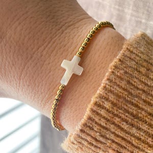 May include: Gold beaded bracelet featuring a white cross pendant. The bracelet is made of small, round gold beads and has a delicate appearance. The cross is a light, pearly white color.