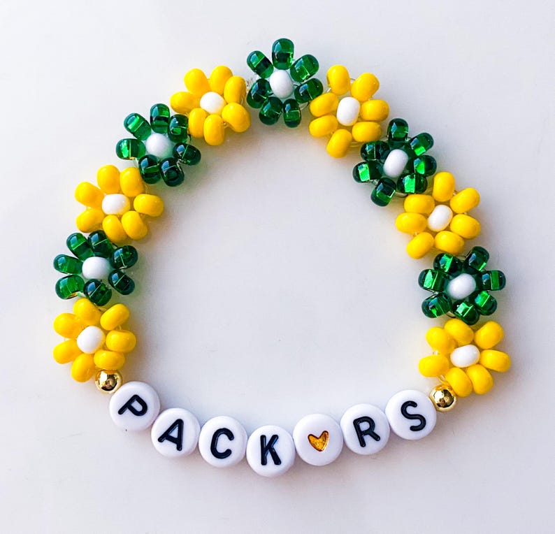May include: A beaded bracelet featuring yellow and green flower designs. The bracelet has white beads with the word "PACKERS" spelled out. The bracelet is a vibrant accessory with a playful floral theme.