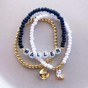 K&ouml;nnte beinhalten: Drei Armb&auml;nder mit Perlen. Ein Armband hat den Namen "WALLEN" in wei&szlig;en Buchstabenperlen. Die anderen Armb&auml;nder sind goldfarben, schwarz und wei&szlig;. Zu den Anh&auml;ngern geh&ouml;ren ein goldener Cowboyhut und ein wei&szlig;er und goldener Cowboy-Stiefel.