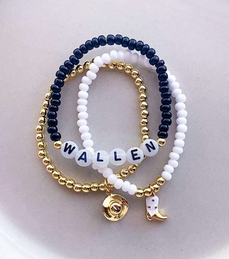 K&ouml;nnte beinhalten: Drei Armb&auml;nder mit Perlen. Ein Armband hat den Namen "WALLEN" in wei&szlig;en Buchstabenperlen. Die anderen Armb&auml;nder sind goldfarben, schwarz und wei&szlig;. Zu den Anh&auml;ngern geh&ouml;ren ein goldener Cowboyhut und ein wei&szlig;er und goldener Cowboy-Stiefel.