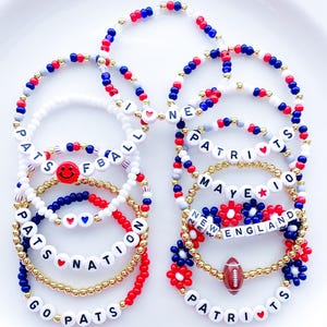 May include: A collection of beaded bracelets in red, white, blue, and gold. Some bracelets spell out "PATRIOTS", "GO PATS", "NATION", and "NEW ENGLAND". Several bracelets feature heart and football charms, and smiley face beads.