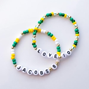 May include: Two beaded bracelets with white letter beads spelling "LOVE 10" and "JACOBS 8". The bracelets feature green, yellow, white, and gold beads. The bracelets are on a white background.