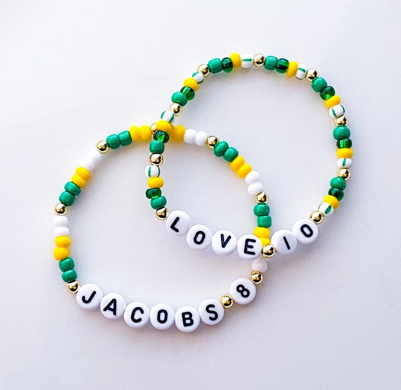 May include: Two beaded bracelets with white letter beads spelling "LOVE 10" and "JACOBS 8". The bracelets feature green, yellow, white, and gold beads. The bracelets are on a white background.