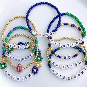 May include: Several beaded bracelets in gold, green, and blue. Some bracelets spell out "GO IRISH", "NOTRE DAME", and "FIGHTING". One bracelet features a small football charm. The bracelets are on a white surface.