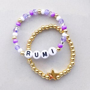 Pode incluir: Duas pulseiras de contas. Uma apresenta contas douradas com um berloque de estrela dourada. A outra tem contas roxas, brancas e transparentes, e contas de letras brancas que soletram "RUMI".