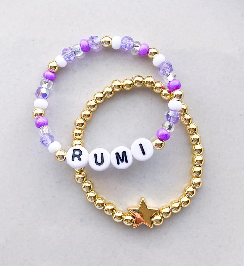 Pode incluir: Duas pulseiras de contas. Uma apresenta contas douradas com um berloque de estrela dourada. A outra tem contas roxas, brancas e transparentes, e contas de letras brancas que soletram "RUMI".