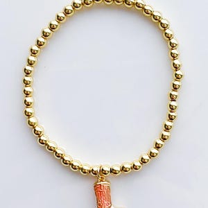 Puede incluir: Pulsera de cuentas doradas con un dije de una peque&ntilde;a bota de vaquero detallada en tonos rojos y dorados. La pulsera presenta peque&ntilde;as cuentas redondas de color dorado ensartadas. El dije de la bota cuelga del centro de la pulsera.