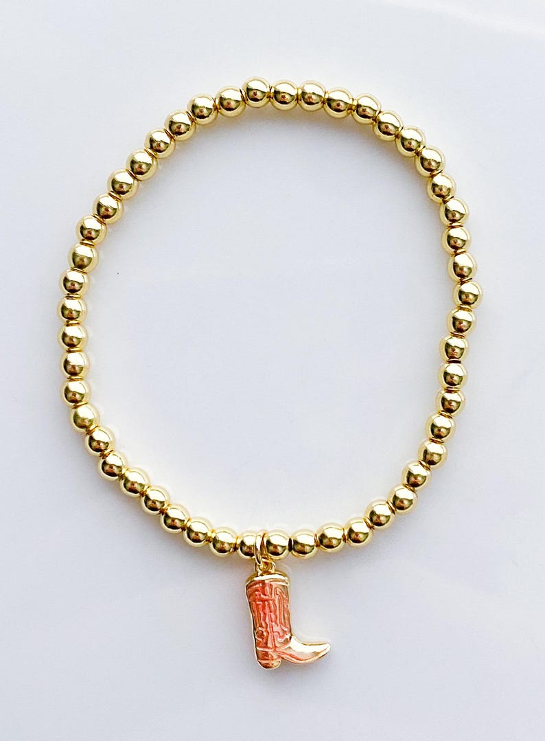 Puede incluir: Pulsera de cuentas doradas con un dije de una peque&ntilde;a bota de vaquero detallada en tonos rojos y dorados. La pulsera presenta peque&ntilde;as cuentas redondas de color dorado ensartadas. El dije de la bota cuelga del centro de la pulsera.