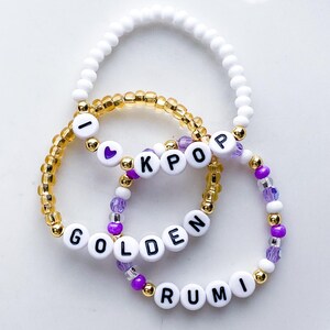 Pode incluir: Tr&ecirc;s pulseiras de contas. Uma &eacute; branca, outra dourada e a terceira roxa e branca. A pulseira dourada tem as letras "I KPOP". A pulseira roxa e branca tem as palavras "GOLDEN RUMI".
