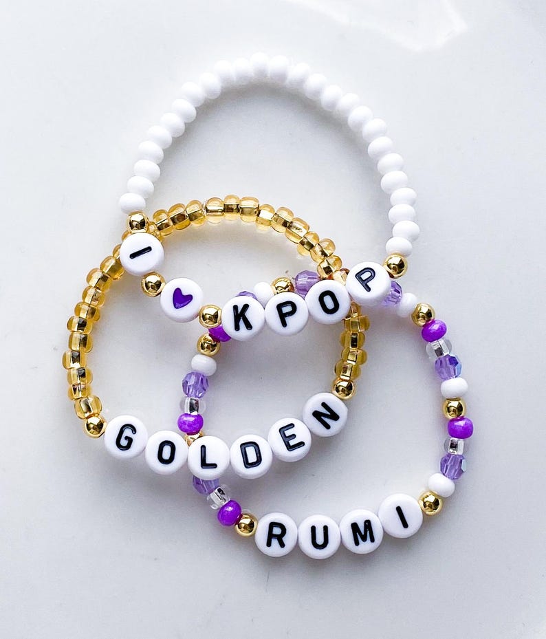 Pode incluir: Tr&ecirc;s pulseiras de contas. Uma &eacute; branca, outra dourada e a terceira roxa e branca. A pulseira dourada tem as letras "I KPOP". A pulseira roxa e branca tem as palavras "GOLDEN RUMI".