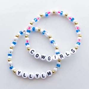 Puede incluir: Dos pulseras de cuentas. Una pulsera deletrea "COWGIRLS" en cuentas de letras blancas. La otra pulsera deletrea "YLLYLM". Las pulseras tienen cuentas blancas, doradas, azules y rosas.