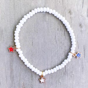 May include: White beaded bracelet with three star charms in red, white, and blue. The bracelet is made of small, round white beads and has gold-colored metal accents. The star charms are in the colors of the American flag.