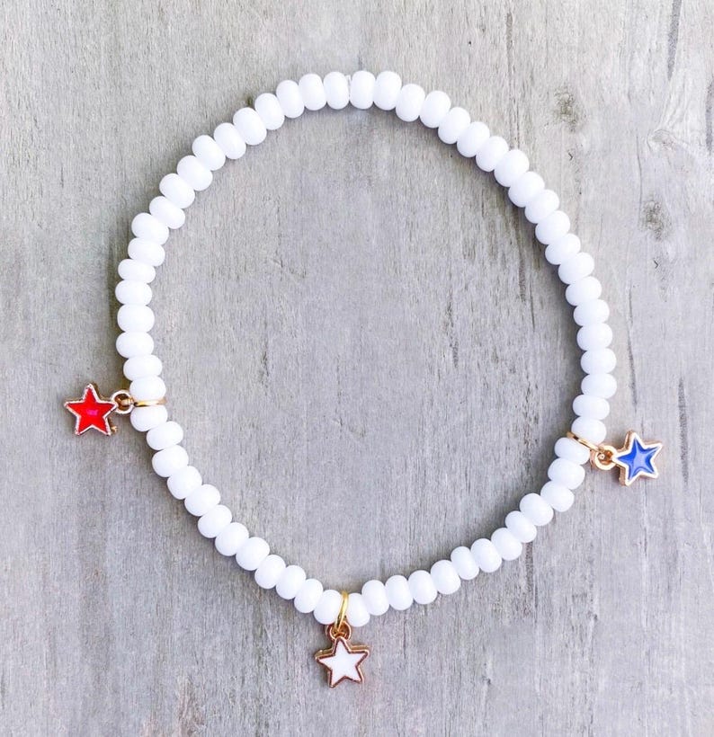 May include: White beaded bracelet with three star charms in red, white, and blue. The bracelet is made of small, round white beads and has gold-colored metal accents. The star charms are in the colors of the American flag.