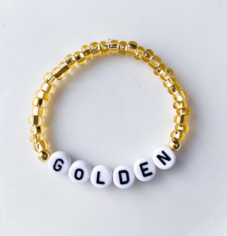 Pode incluir: Uma pulseira de contas com a palavra "GOLDEN" escrita em letras pretas sobre contas brancas. O resto da pulseira &eacute; feito de pequenas contas douradas. A pulseira &eacute; circular e est&aacute; sobre uma superf&iacute;cie branca.