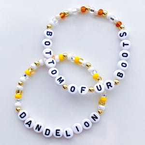 Puede incluir: Dos pulseras de cuentas con letras blancas que deletrean "BOTTOM OF FUR" y "DANDELION BOOTS". Las pulseras tienen cuentas doradas, amarillas y transparentes. Las pulseras est&aacute;n sobre una superficie blanca.