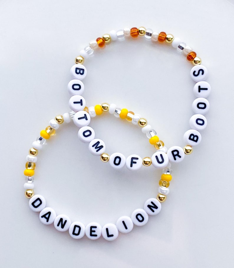 Puede incluir: Dos pulseras de cuentas con letras blancas que deletrean "BOTTOM OF FUR" y "DANDELION BOOTS". Las pulseras tienen cuentas doradas, amarillas y transparentes. Las pulseras est&aacute;n sobre una superficie blanca.