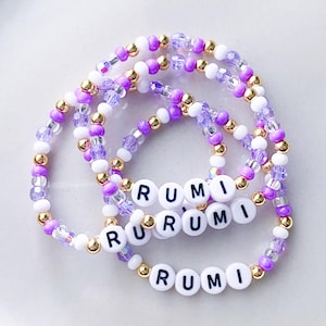 Pode incluir: Tr&ecirc;s pulseiras de contas com o nome "RUMI" escrito com contas de letras brancas. As pulseiras combinam contas brancas, roxas e douradas. As pulseiras est&atilde;o em uma superf&iacute;cie branca.