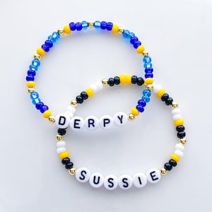 Pode incluir: Duas pulseiras de contas. Uma pulseira diz "DERPY" com contas de letras brancas. A outra pulseira diz "SUSSIE". Ambas as pulseiras t&ecirc;m contas azuis, amarelas, pretas, brancas e douradas.