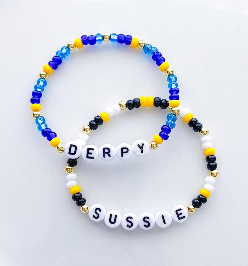 Pode incluir: Duas pulseiras de contas. Uma pulseira diz "DERPY" com contas de letras brancas. A outra pulseira diz "SUSSIE". Ambas as pulseiras t&ecirc;m contas azuis, amarelas, pretas, brancas e douradas.