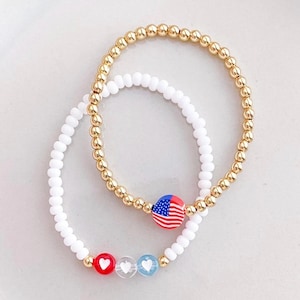May include: Two beaded bracelets. One is gold-colored with small round beads, and the other is white. One bracelet has a charm with the American flag, and the other has red, clear, and blue heart charms.