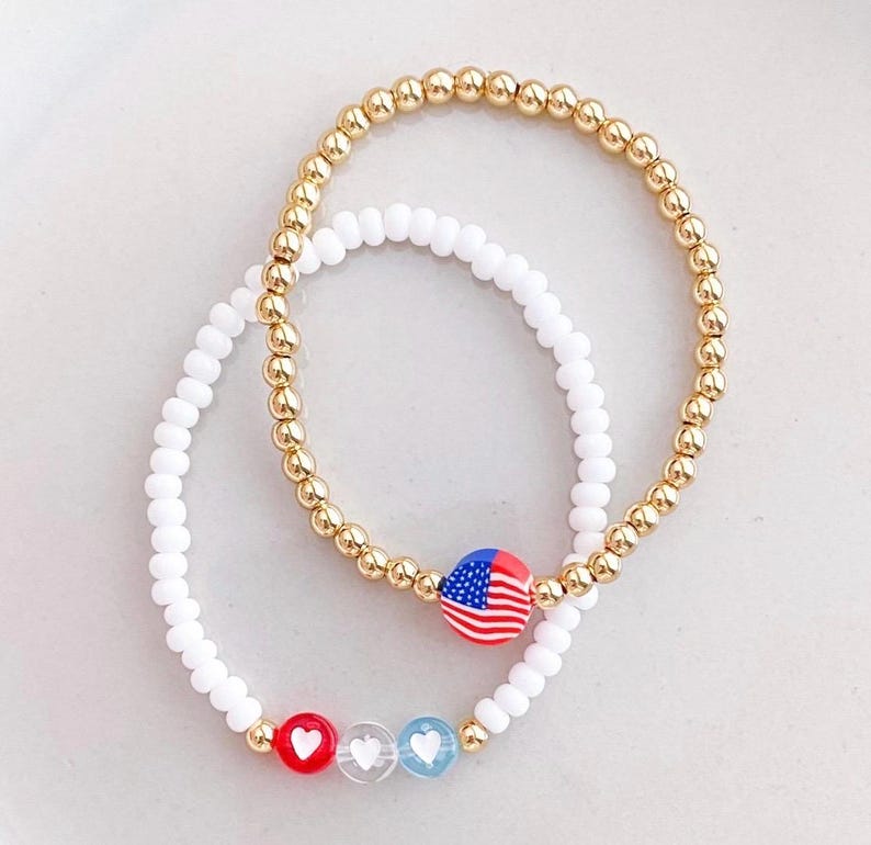 May include: Two beaded bracelets. One is gold-colored with small round beads, and the other is white. One bracelet has a charm with the American flag, and the other has red, clear, and blue heart charms.