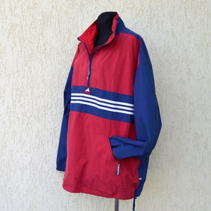 Op de afbeelding: Een rood en blauw windjack met een wit Adidas-logo en drie witte strepen op de borst. Het jack heeft een halve rits en een trekkoord aan de onderkant.