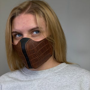 Puede incluir: Una mascarilla facial de cuero marrón con ribete negro y estampado de cocodrilo. La mascarilla tiene pequeños agujeros para la transpirabilidad.