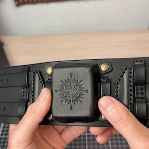 Puede incluir: Cinturón de cuero negro con un diseño de rosa de los vientos en una hebilla cuadrada. El cinturón tiene múltiples presillas. El fondo muestra herramientas de marroquinería.