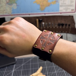 May include: A brown leather cuff bracelet with a Celtic knot design and brass rivets. The bracelet is approximately 2 inches wide and is fastened with a buckle. The design is embossed into the leather.