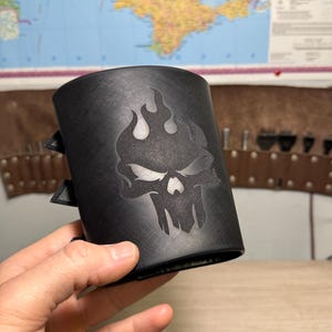 Puede incluir: Brazalete de cuero negro con diseño de calavera y llamas. El brazalete tiene dos correas y hebillas ajustables. La imagen muestra el brazalete contra un mapa del mundo.